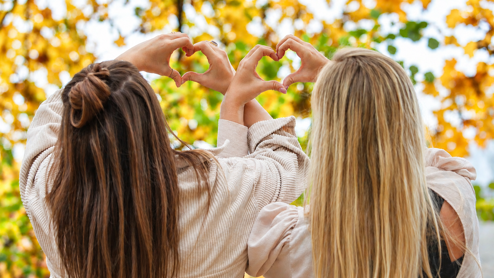 Zwei Frauen bilden mit ihren Händen Herzen vor herbstlichen Bäumen.