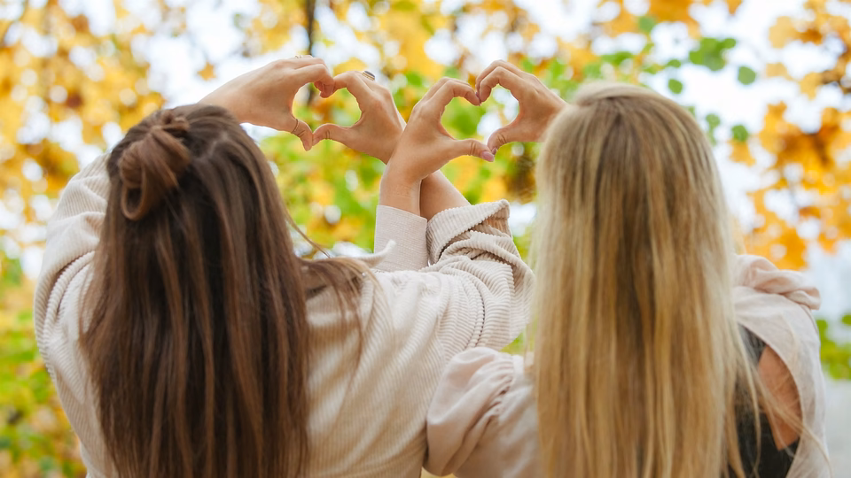 Silvia und Kristina formen mit ihren Händen Herzen vor herbstlichen Bäumen.