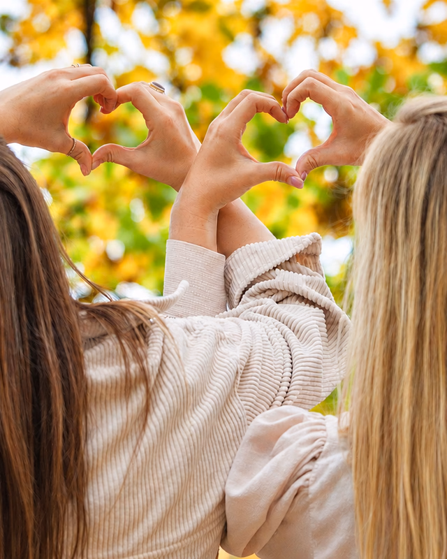Silvia und Kristina formen mit ihren Händen Herzen vor herbstlichen Bäumen.